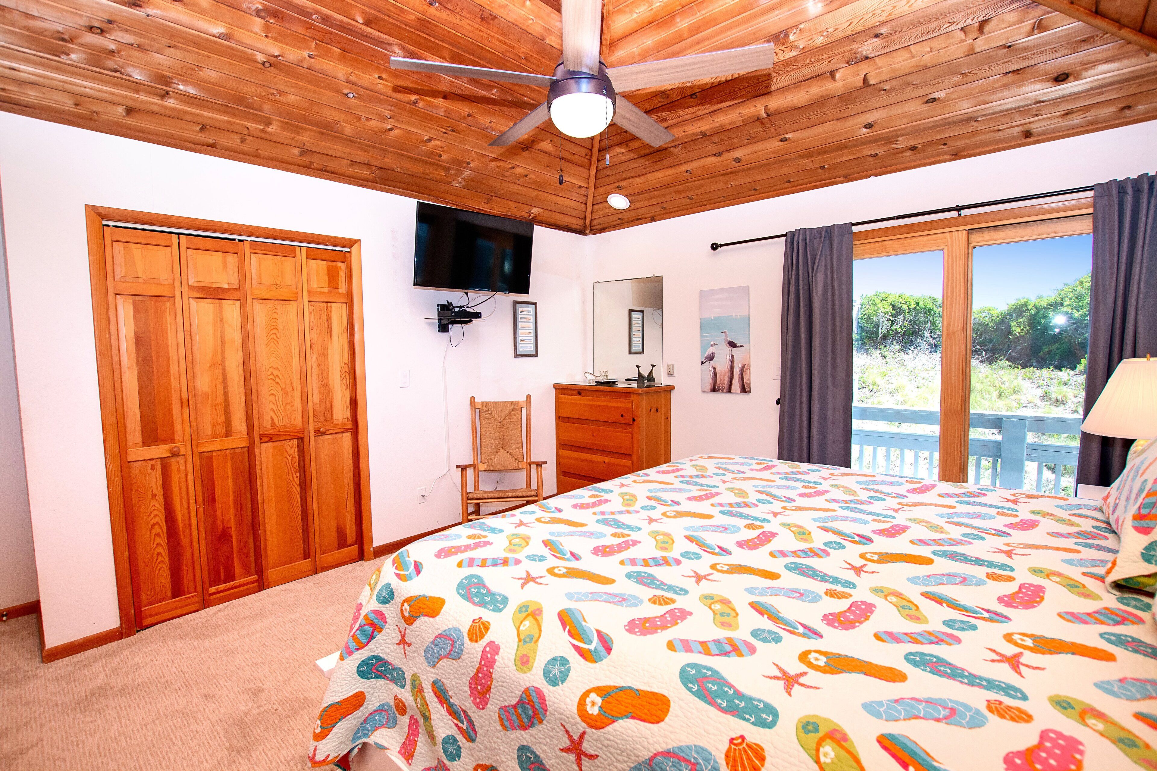 Une chambre avec un plafond en bois, un lit recouvert d’une courtepointe aux couleurs vives, une télévision, une commode et une fenêtre avec des rideaux.