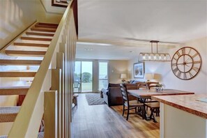 Stairs lead to a spacious dining area with a table, chairs, and a large clock, offering a view through a glass door to an outdoor space.