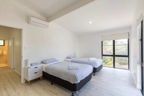 Two single beds with matching headboards and footboards, a bedside table, and a lamp create a cozy space with a large window offering a view of the outdoors.