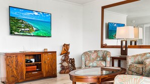 Una sala de estar con un aparador de madera, una mesa de centro redonda de vidrio, sillones con estampado, un televisor de pantalla plana grande y un espejo.