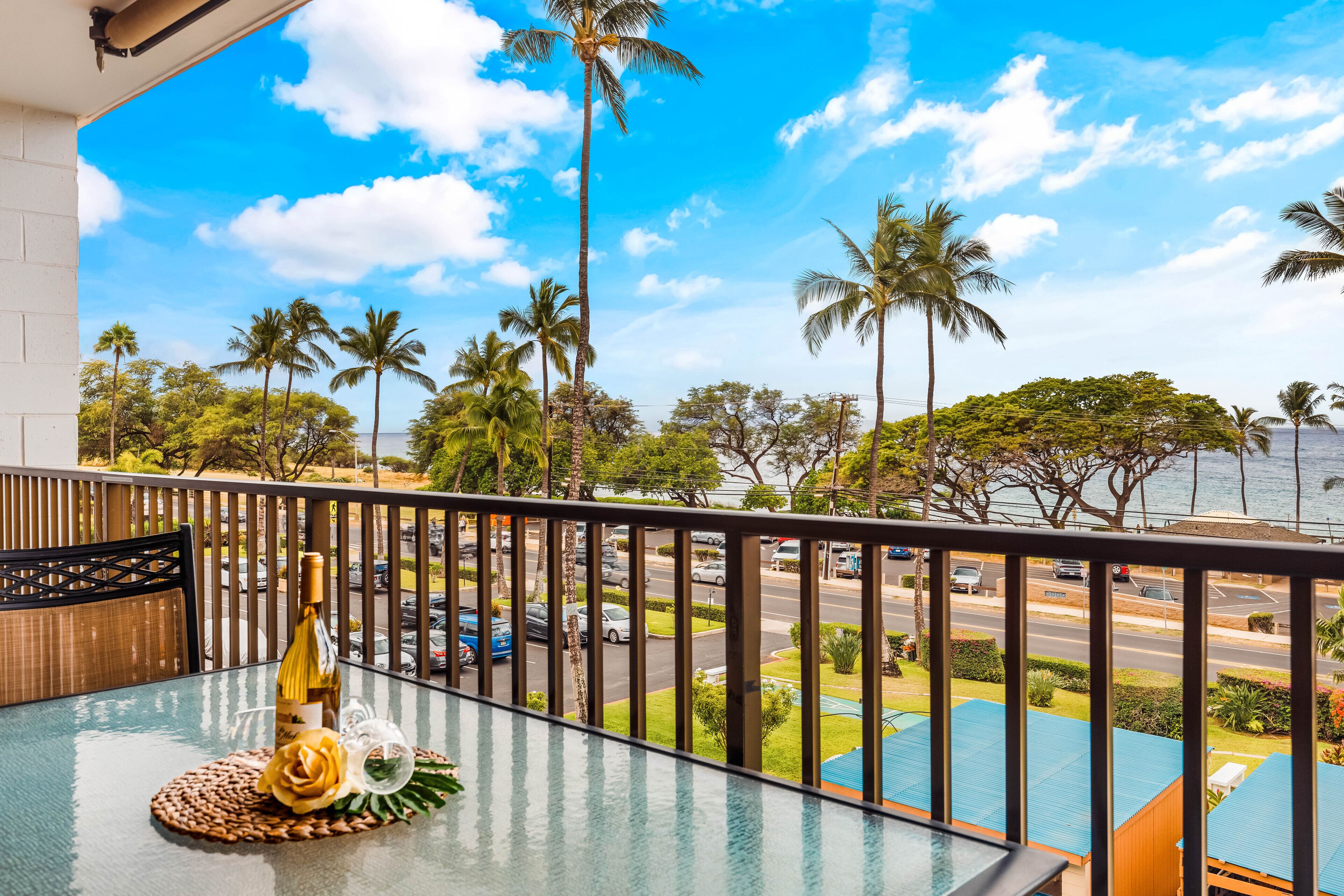 A balcony with a glass table, a bottle, and a decorative item. It overlooks a street with cars and palm trees.