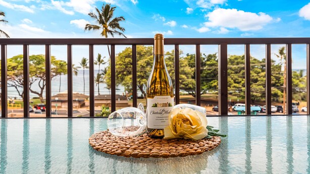 Una botella de vino Chateau Moka, una copa y una rosa amarilla sobre una mesa de vidrio con vistas a palmeras y un cielo despejado.