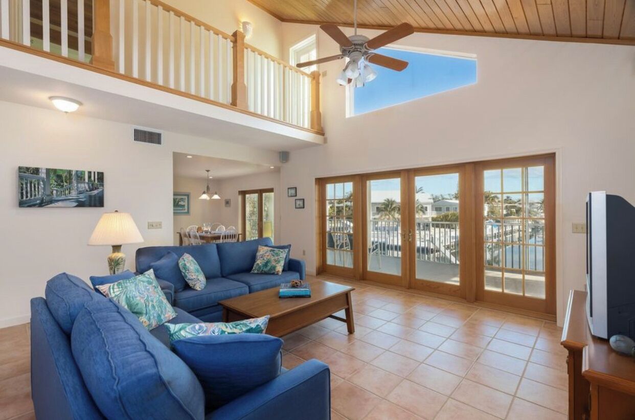 A spacious living room with a blue sofa set, a wooden coffee table, and a ceiling fan. Large windows offer a view of a balcony with outdoor furniture.