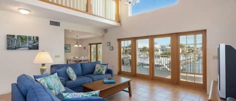 A spacious living room with a blue sofa set, a wooden coffee table, and a ceiling fan. Large windows offer a view of a balcony with outdoor furniture.