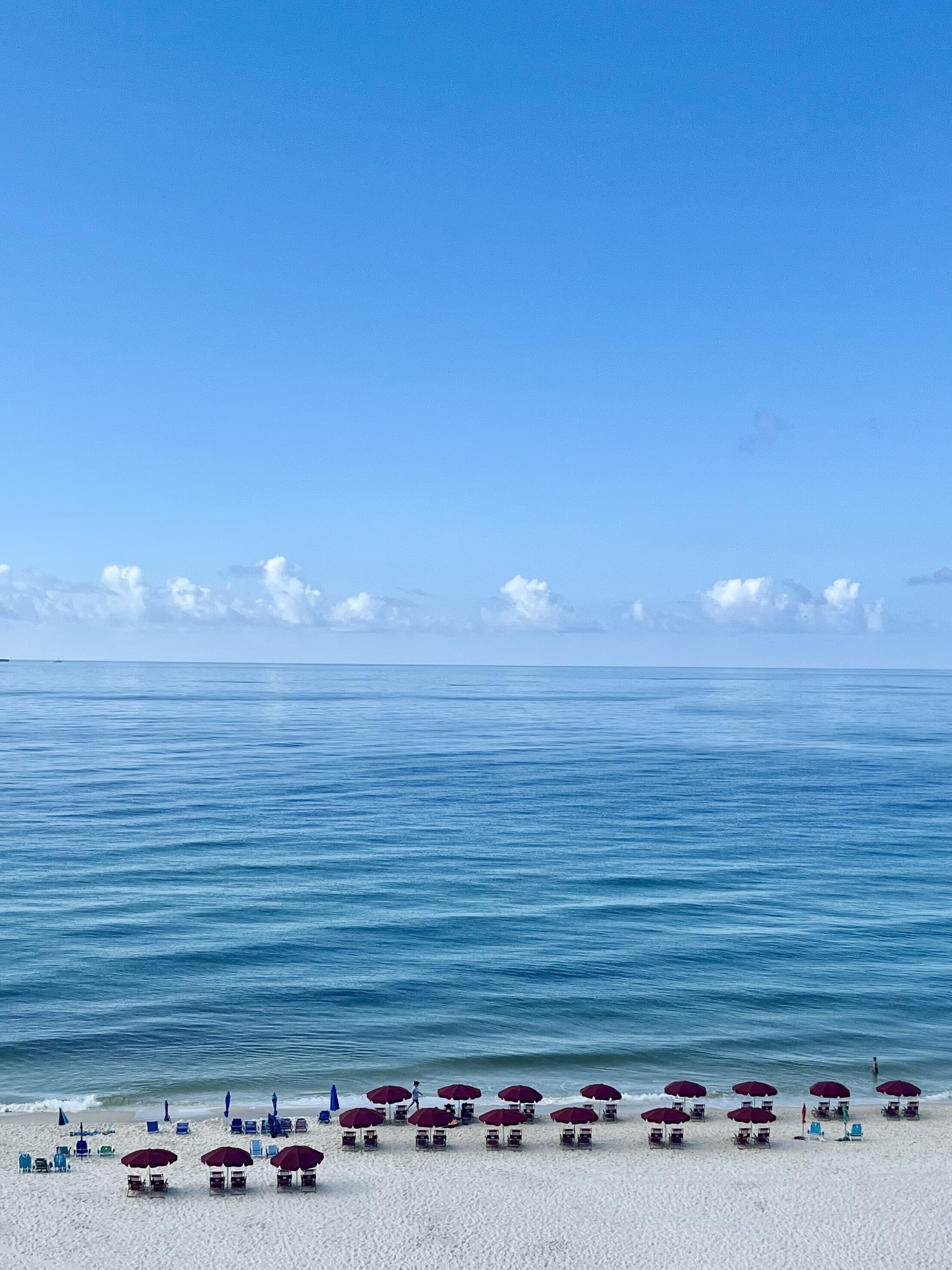 Une plage de sable blanc, sous un ciel bleu azur. De nombreuses rangées de chaises de plage et de parasols bordent le rivage.