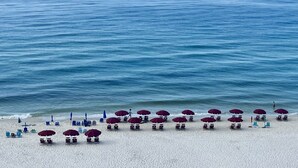 Una spiaggia con sabbia bianca e un cielo azzurro limpido. Si vedono file di sedie a sdraio e ombrelloni lungo la battigia.