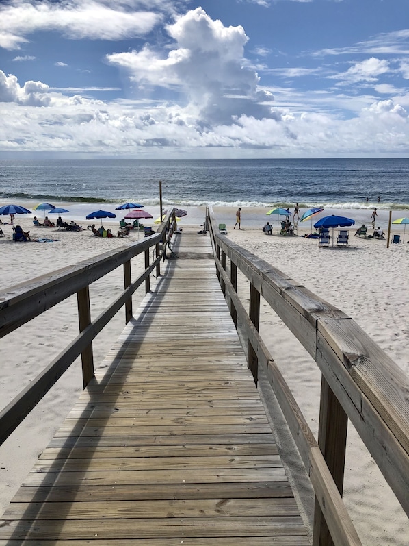 Ein hölzerner Steg führt zu einem Strand mit Sonnenschirmen und Liegestühlen.