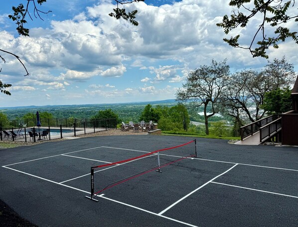 Una cancha de tenis con red, rodeada por una cerca, con vistas a un paisaje pintoresco con árboles y una lejana vista de un lago.