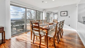 Een ruime eetkamer met een grote houten tafel en stoelen, grote ramen met uitzicht op het landschap, en een moderne keukenhoek.