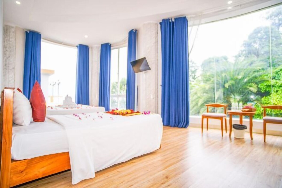 A hotel room with a large bed, a small table, and a chair. There are blue curtains and a large window with a view of greenery outside.