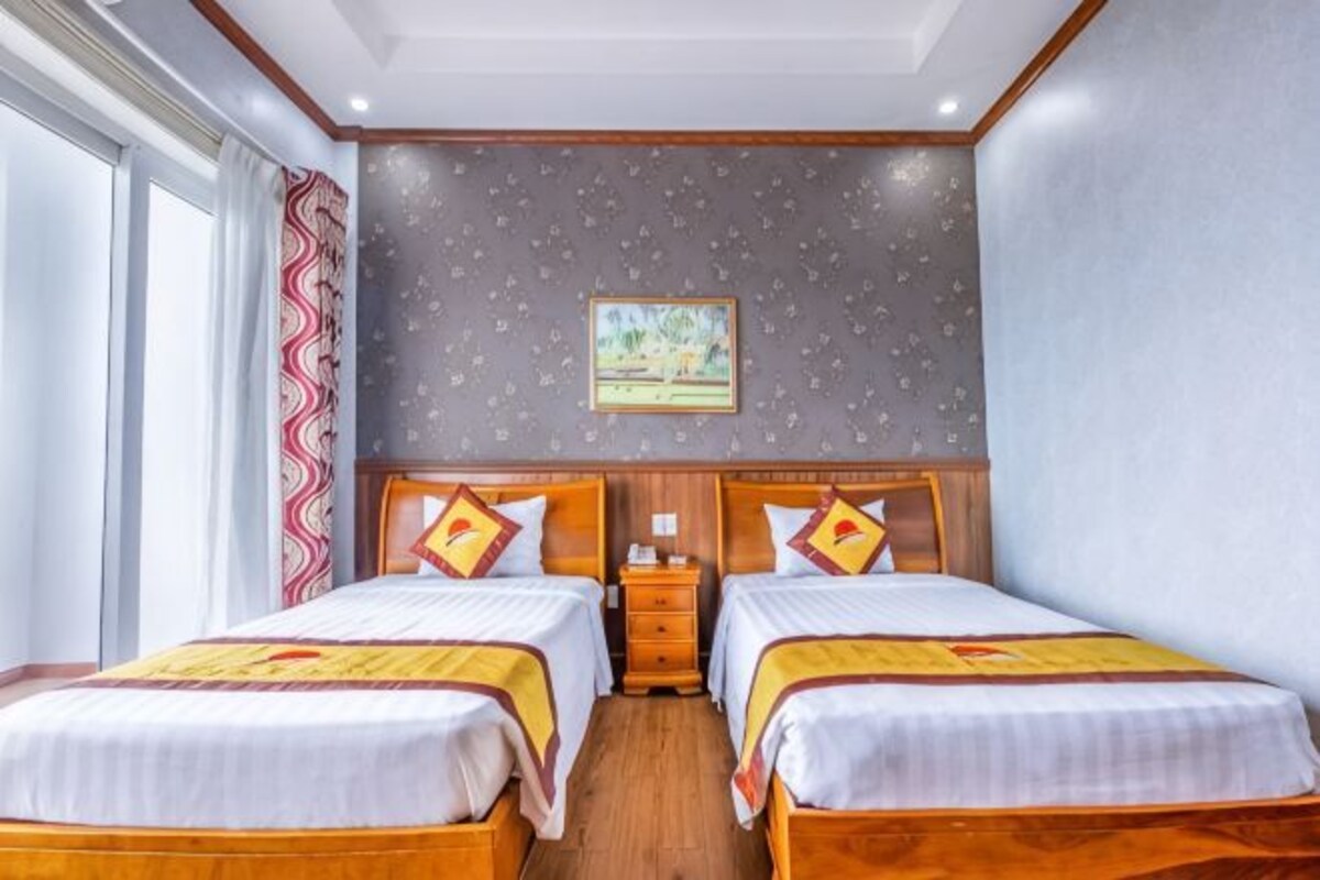 A hotel room with two beds, a nightstand, a painting on the wall, and a window with curtains.