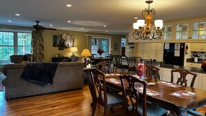 A spacious living room with a large sofa, dining table, and chairs, a chandelier, and a kitchen with white cabinets.