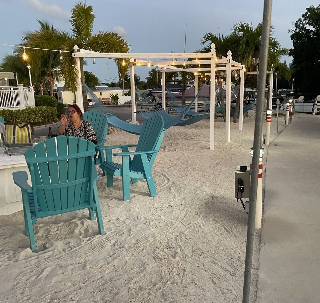 Un espacio de descanso frente a la playa con sillas Adirondack turquesa y una pérgola blanca con guirnaldas de luces.