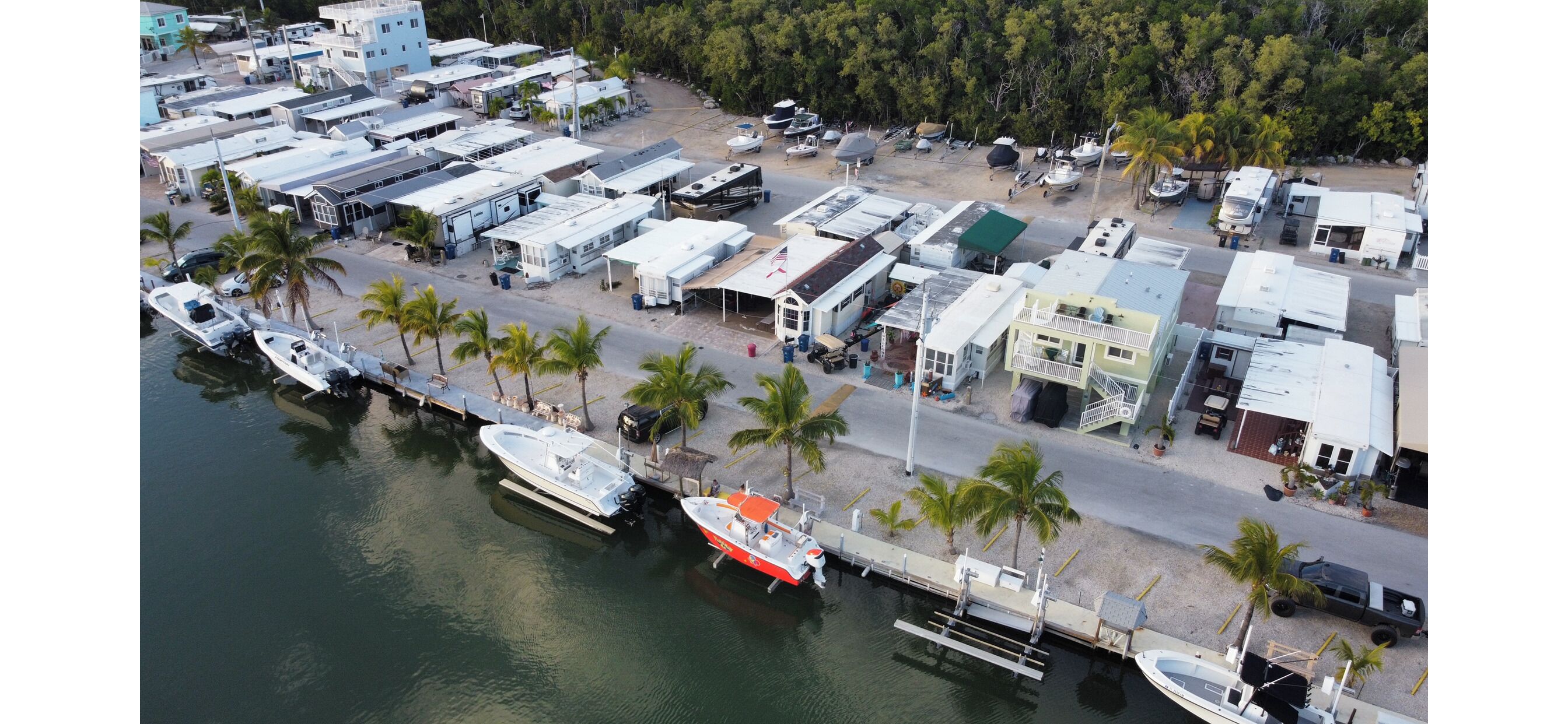 An aerial view of a marina with boats docked, a road lined with buildings, and palm trees.