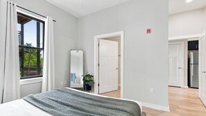A bedroom with a bed, a window with curtains, a door, and a hallway leading to a kitchen.