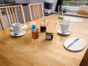 Une table de salle à manger en bois, avec des tasses et des soucoupes blanches, et un verre d’eau. On y trouve également deux pots de confiture et une bouteille de liquide vaisselle.