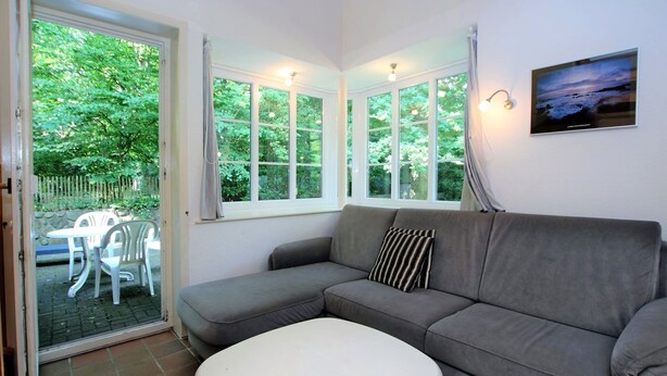 Una habitación con un sofá gris, un otoman blanco y una mesa de comedor con sillas. Hay ventanas grandes con cortinas, y un cuadro en la pared.