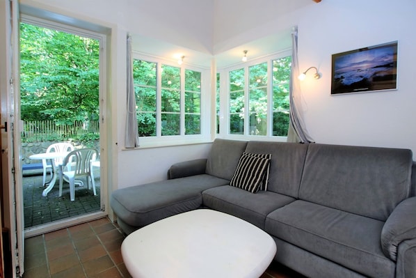 A room with a grey sofa, a white ottoman, and a dining table with chairs. There are large windows with curtains, and a picture on the wall.