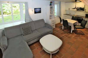 A living area with a sofa, a small table, a kitchen with a dining table and chairs, and a window with curtains. - Gmelinstraße 19 d - Hausscheibe mit 2- Zimmern auf ca. 52m² und Terrasse (Wyk auf Föhr)
