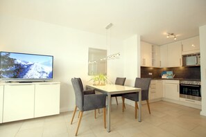 A modern kitchen with white cabinets, a dining table with four chairs, a flat-screen TV, and a built-in oven.