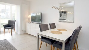 A modern dining area with a white table, grey upholstered chairs, a flat-screen TV on a white cabinet, and a large mirror on the wall.