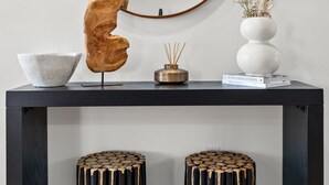 Una habitación moderna con un mueble consola de madera oscura, un espejo redondo grande, un jarrón blanco con flores secas, un cuenco blanco y dos taburetes únicos con un diseño de pintura goteando.