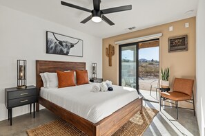 A bedroom with a wooden bed, a ceiling fan, a nightstand, a chair, and a large window with a view of the desert.