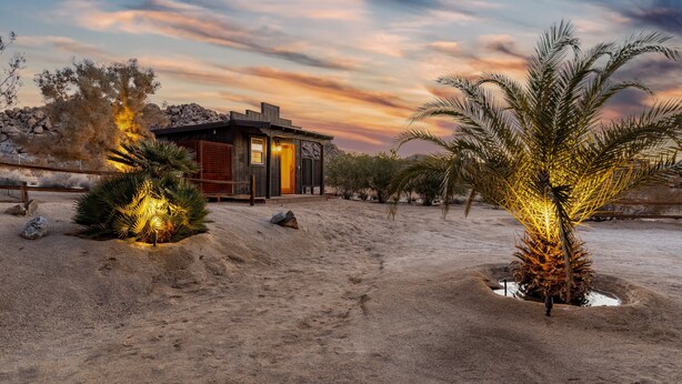 Paisaje desértico con una casa moderna, revestida de madera y con luz cálida en su interior.
