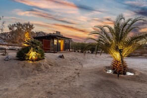 Paisaje desértico con una casa moderna, revestida de madera y con luz cálida en su interior.
