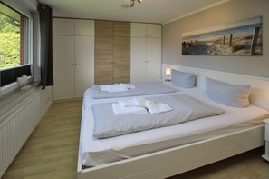 A bedroom with two beds, a window with blinds, a radiator, a painting on the wall, and a bedside table with a lamp.