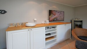 A kitchen with white cabinets, a wooden countertop, a television mounted on the wall, and a wicker chair. - Schmalstieg 11 a, Wyker Perle - Holiday home with 3 rooms on approx. 81m² (Wyk auf Föhr)