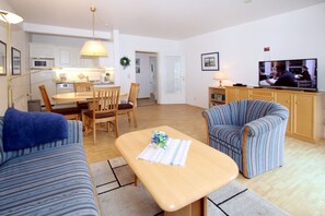 A living room with a sofa, armchair, coffee table, and a television. There is a dining area with a table and chairs.
