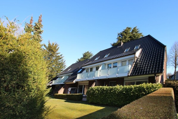 A two-story residential building with a dark tiled roof, multiple windows, and a well-maintained garden with hedges.