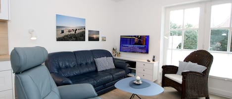 Un soggiorno moderno con divano, poltrona e sedia in vimini, televisore e un tavolino con un oggetto decorativo.