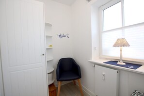 A room with a white door, a black chair, a window with blinds, a radiator, and a wall-mounted lamp. - Olhörnstieg 6, Whg. 2 Inselliebe - Apartment with 3 rooms on approx. 53m² (Wyk auf Föhr)