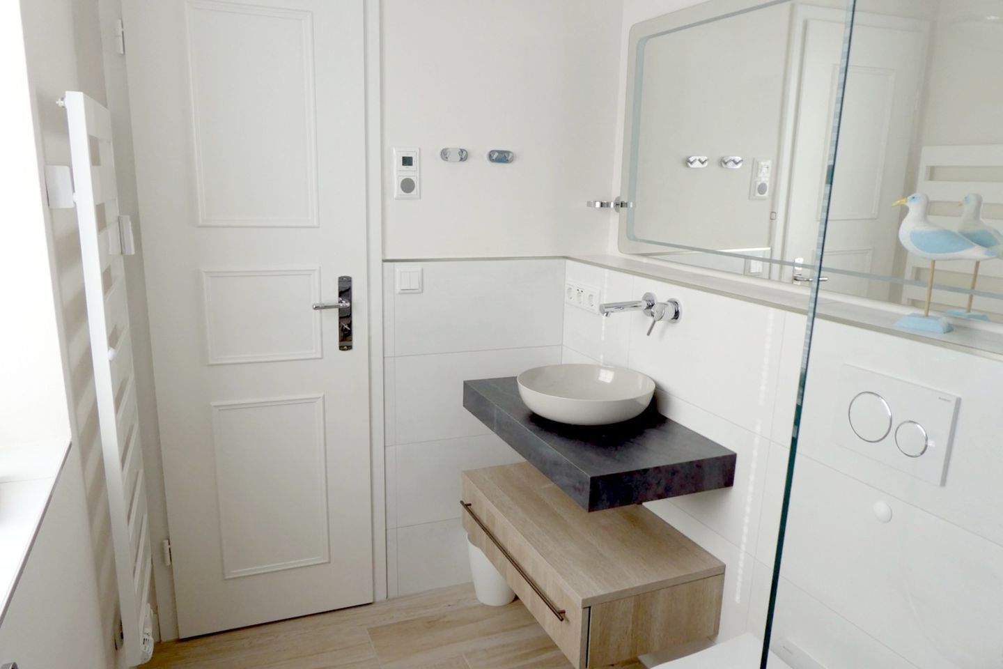 Un baño moderno con una puerta blanca, cabina de ducha de vidrio, lavamanos blanco con encimera negra y un banco de madera.