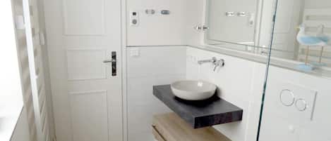 A modern bathroom with a white door, a glass shower enclosure, a white sink with a black countertop, and a wooden bench.