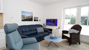A modern living room with a sofa, armchair, and wicker chair, a television, and a small table with a decorative item. - Olhörnstieg 6, Whg. 2 Inselliebe - Apartment with 3 rooms on approx. 53m² (Wyk auf Föhr)