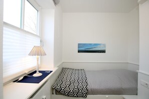 A small, well-lit room with a bed, a desk with a lamp, and a window with blinds. - Olhörnstieg 6, Whg. 2 Inselliebe - Apartment with 3 rooms on approx. 53m² (Wyk auf Föhr)