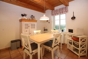 Una cocina con una mesa de madera, sillas blancas, una lámpara colgante, un ventanal con un adorno de cuadros y un microondas blanco sobre una estantería.