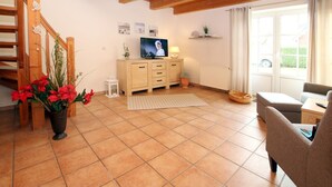 Un salon avec un escalier en bois, un téléviseur posé sur un meuble en bois, une plante en pot et une fenêtre avec des rideaux.