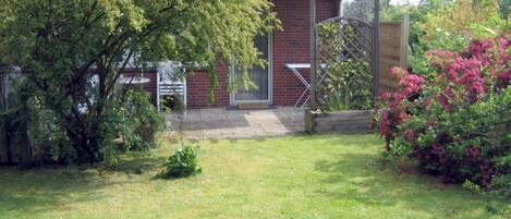 A brick building with a metal roof, a wooden fence, and a garden with grass and shrubs.