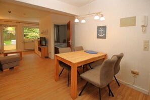 A room with a wooden dining table, two chairs, a bench, a television, and a kitchen area.