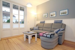 A living room with a grey sofa, a small white table, and a round ottoman. There are three framed pictures on the wall, and a window with blinds.