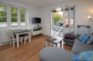 A modern, well-lit room with a dining table, a sofa, a TV, and a view of the outside.