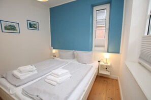 A double bed with white linens, two bedside tables with lamps, a window with blinds, and two framed pictures on the wall.