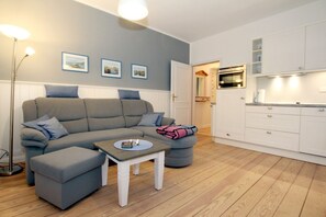 A modern living room with a grey sofa, a small table with a book and candles, a kitchen area with white cabinets, and framed pictures on the wall.