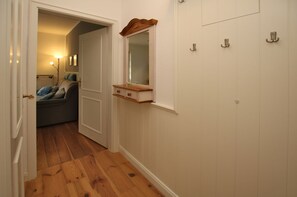 A hallway with wooden flooring, a door leading to a room with a sofa, and a mirror on the wall.