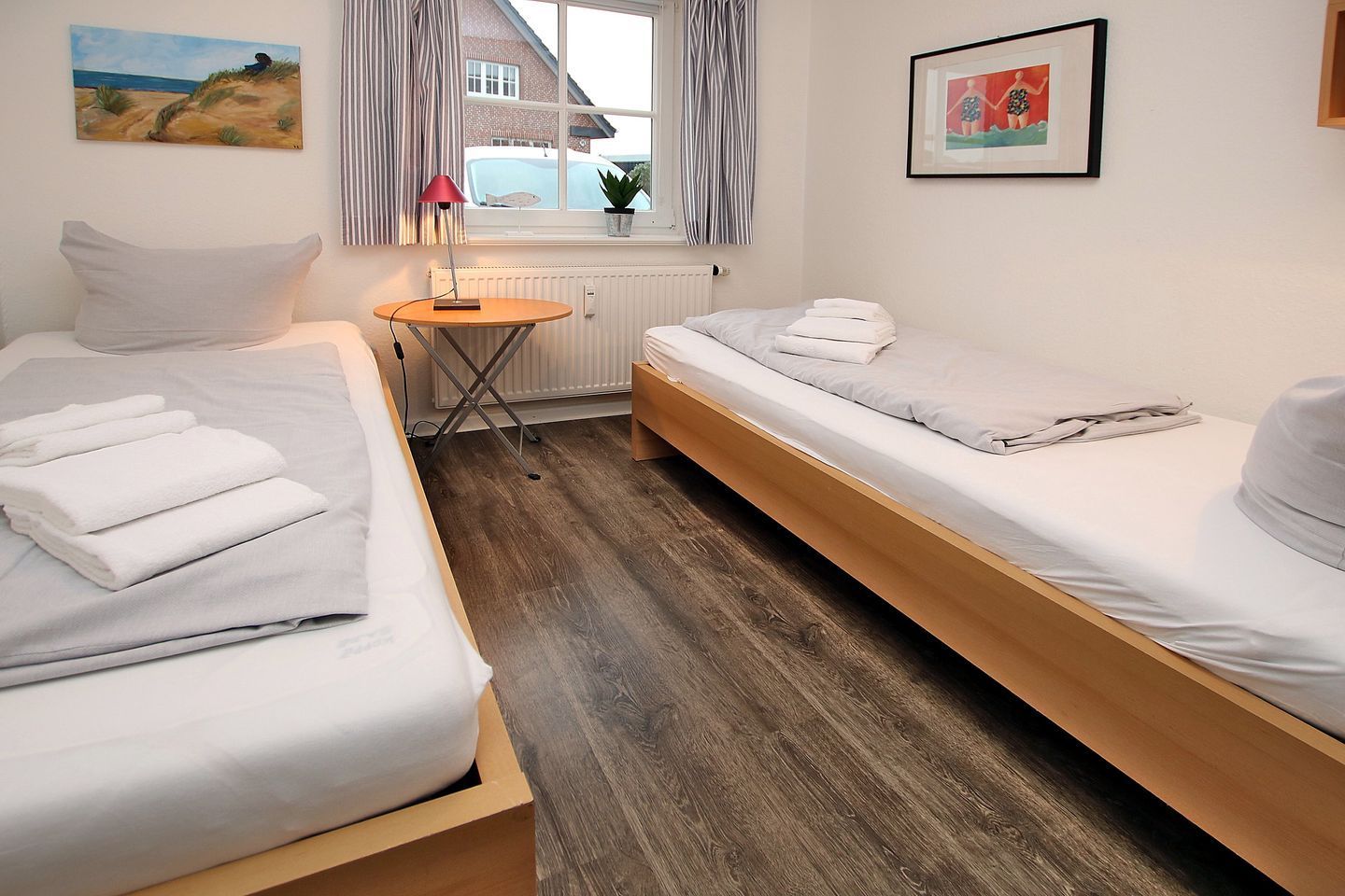 Quarto com duas camas individuais, uma mesinha com abajur, radiador e janela com cortinas.