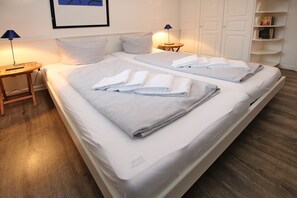 A double bed with white linens and folded towels, a bedside table with a lamp, a small round table with a lamp, and a bookshelf with books. - Badestraße 18a, Whg. 1 - vacation apartment with 3 rooms, approx. 60m², in the center of Wyk, (Wyk auf Föhr)